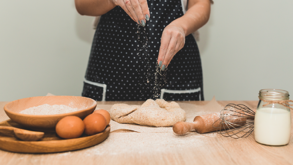 Person sprinkling flour over dough on a kitchen table with simple baking ingredients nearby
