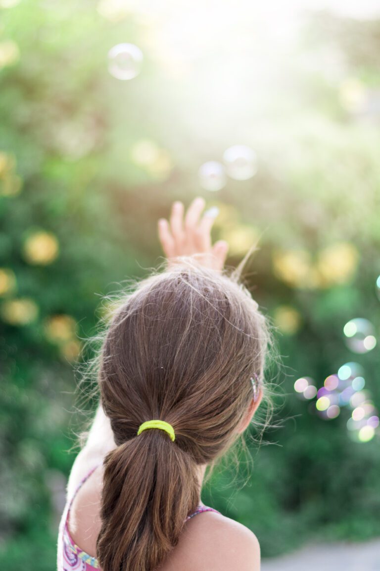 Little girl with a ponytail raising her hand outdoors with soap bubbles in soft sunlight, green bokeh background